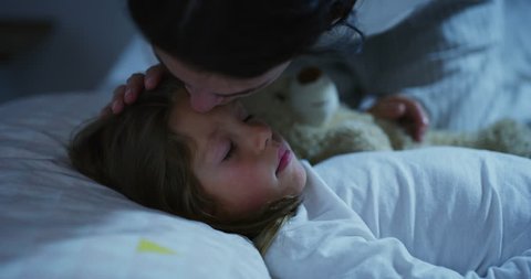 Portrait of a woman (girl) while she is kissing her daughter sleeping in the bed with the teddy bear. Concept: Family, love, dreams