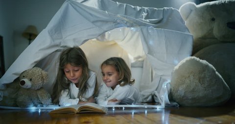 Two little girls sisters (kids) read stories in the dark under the blanket illuminating with a torch. Concept: Love, Family, Dreams