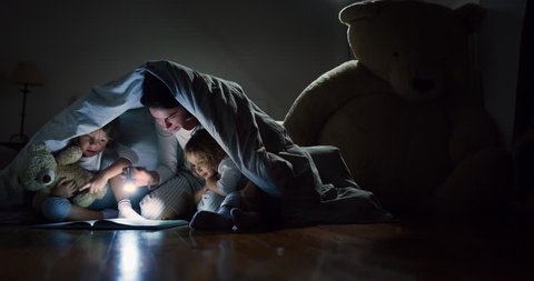 Slow motion of young mother is reading a goodnight story to her little daughters in the dark illuminating with a torch under the blanket. Concept of family, childhood, love, health, education
