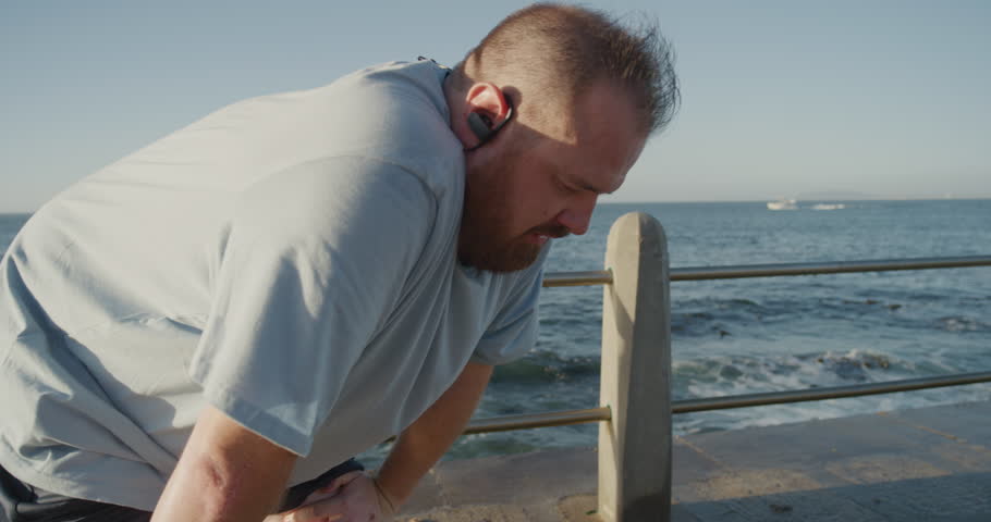 young overweight runner man resting exhausted after intense running exercise training intense weight loss challenge on seaside morning listening to music at sunrise close up