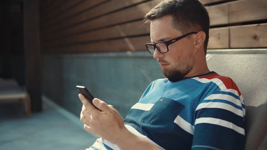 Portrait of a man in glasses with a smartphone, man sitting online and chatting, texting sms on cell. Man sunbathing in a t-shirt.