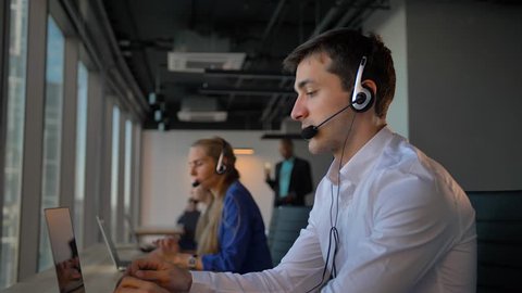 Attractive business man in headset calling to cuctomers in business office call center. His female colleagues also calling and communicated by phone with clients. 4k slow motion,