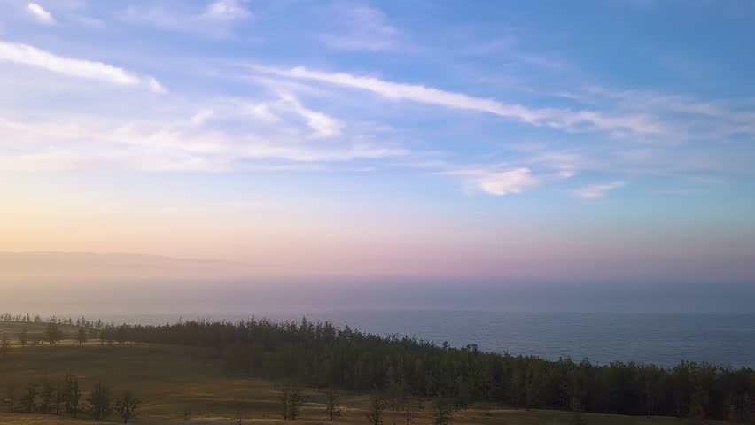 Russia, Lake Baikal, Olkhon Island, sunset over the Small Sea Bay captured from an aerial perspective. The camera reveals a sweeping view of a forested headland silhouetted against the calm waters of the bay, with the sun descending toward the distant horizon. Wispy clouds stretch across a sky that transitions from warm amber and soft gold near the sun to cool blue higher above, reflecting gently on the water surface. The shoreline curves along the frame, dense with dark coniferous trees that contrast with the luminous sky and water. The scene unfolds in smooth, continuous motion, emphasizing the vast scale of the landscape and the tranquil transition from day to evening. The location is known as the Small Sea Bay, a sheltered strait between Olkhon Island and the western shore of Lake Baikal, but no specific structures or named landmarks are visible in this natural, undeveloped view.