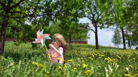 Little cute girl playing with a multicolored pinwheel, a happy child is sitting in dandelions on a spring sunny day.