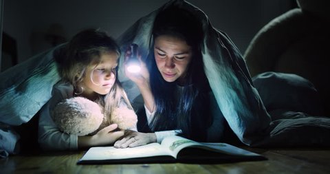 A mother tells stories to her daughter in the dark illuminating with a torch under the blanket. Concept: Love, Family, Dreams