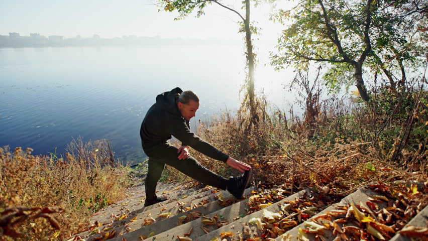 Young guy in sportswear doing warming up exercise while standing on stairs near calm lake on sunny autumn day