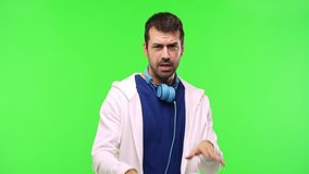 man with sweatshirt and music headphones on green screen chroma key background is a bit nervous and scared stretching hands to the front