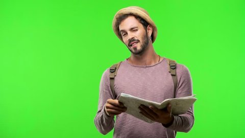 Young traveler man holding a map on green chroma winking, funny joke, symbol of agreement or complicity