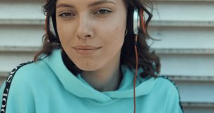 Portrait of beautiful young woman wearing headphones looking to camera and smiling. City urban outdoors. Girl enjoying music and dancing on street. 4K video shooting by handheld gimbal