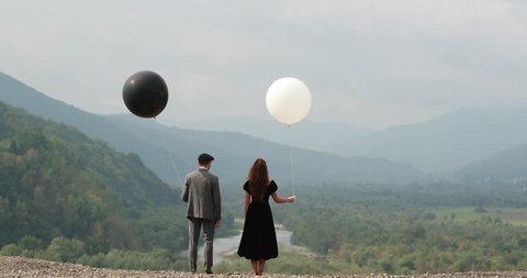 Fly over a couple dressed in dark closes and standing on the rock before beautiful mountain landscape with white and black balloons