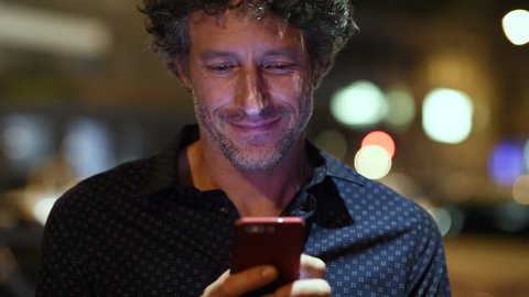 Smiling man using smart phone during night looking at camera. Closeup face of happy mature businessman messaging on cellphone at night on the street with the lights of the road blurred in the backgrou