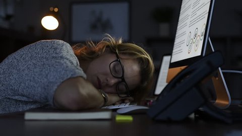 Mature business woman wearing eyeglasses leaning and sleeping on computer keyboard in office. Close up face of tired sleepy businesswoman in dark office.