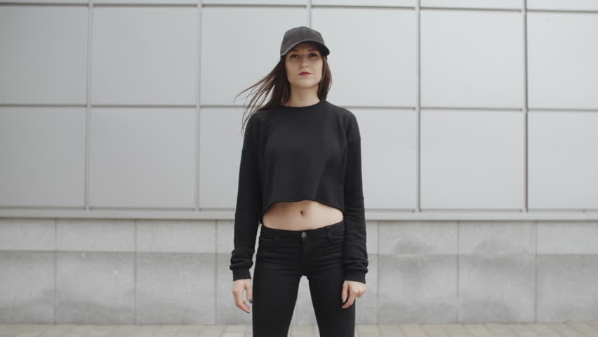 Portrait of brunette woman in black clotihng standing against metal wall of square elements, posing and looking into the camera in the street, urban environment