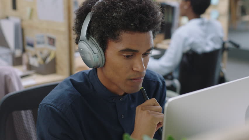 Young mixed race businessman using computer working on business project browsing online listening to music brainstorming ideas wearing headphones in relaxed office workplace