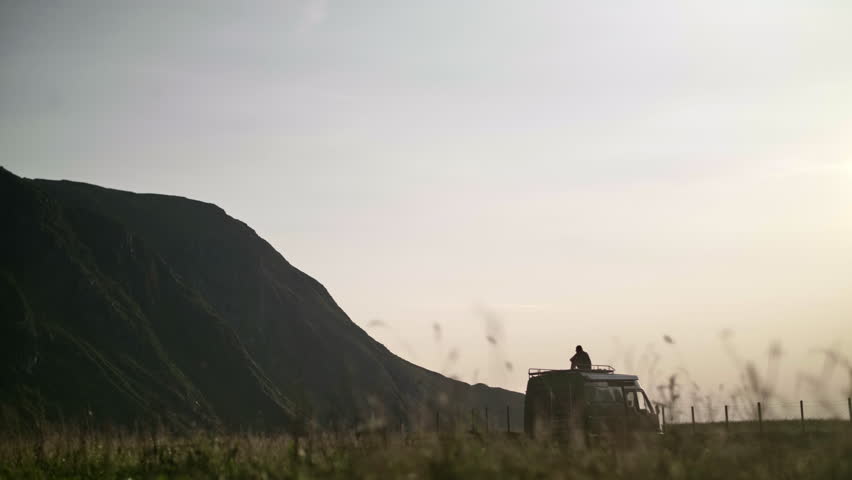 Silhouette of a man sitting on top of a camper van looking at the mountain, along the fields,