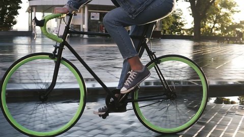 Close up footage of female feet cycling a bicycle in the morning by paved city street with wet asphalt . Side view of a young woman riding a trekking bike with green wheels, wearing sneakers and jeans