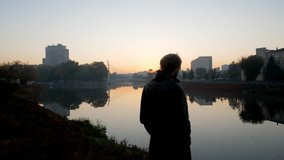 Silhouette of man in black coat standing alone near river and city with sunrise sky