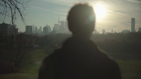 Young Adult Man Stands and Looks at City Skyline from a Distance, Silhouette of Millennial in a Park on Beautiful Sunny Evening at Golden Hour Before Sunset, Cinematic Slow Motion