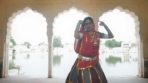 A mid shot of a graceful and attractive dancer in traditional colorful cloths or attire performing in beautiful palace against the lake. An Indian classical or folk dancer dancing in ancient mansion