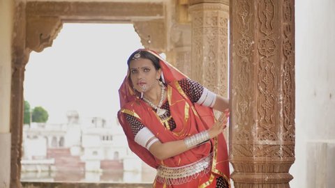 A beautiful and shy woman comes running to the pillar of an old temple in traditional cloths or attire and smiles while looking at the camera. An attractive and Indian ethnic village girl blushing