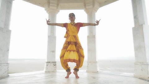 A wide shot of a Indian classical dancer dancing elegantly in white marble temple located on a hilltop. A moving shot of an attractive woman or lady making hand gestures in yellow traditional sari 