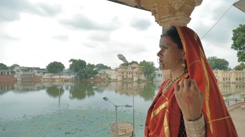 A movement shot of an attractive traditional woman or lady in a colorful sari and jewelry walk out of the house or palace to see the view of a beautiful lake and an old and cultural Indian village. 