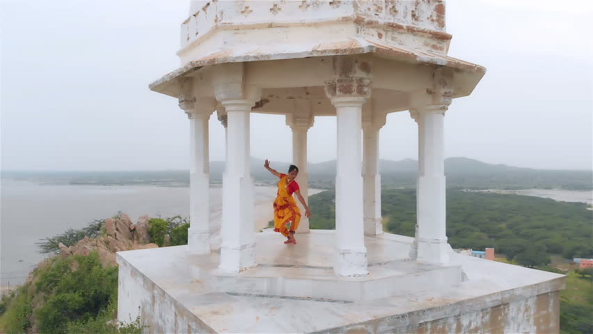  An Aerial shot of an attractive dancer performing in a yellow traditional sari. Movement drone shot of a Classical Indian dancer dancing in marble tomb located on a hilltop surrounded by a desert.