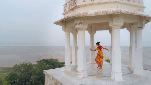 Movement drone shot of a Classical Indian woman dancing in marble tomb located on a hilltop surrounded by a desert. An Aerial shot of a  Bharatanatyam dancer performing in a yellow traditional sari.