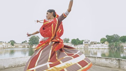 An attractive girl in a traditional attire dancing besides a beautiful lake. A movement shot of young Indian woman in colorful costume performing an Indian classical dance in an old historic village