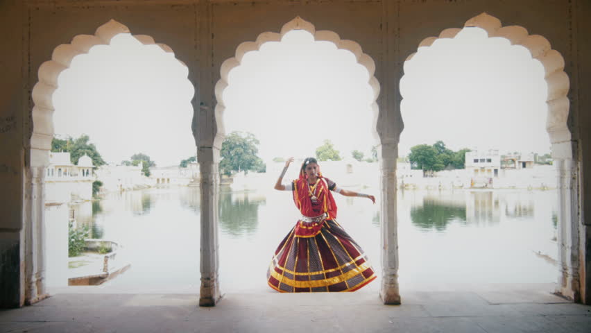 A wide shot of a graceful woman in traditional colorful cloths or attire performing in beautiful palace against the lake. An Indian elegant classical or folk dancer dancing in an ancient mansion | Shutterstock HD Video #1018357840
