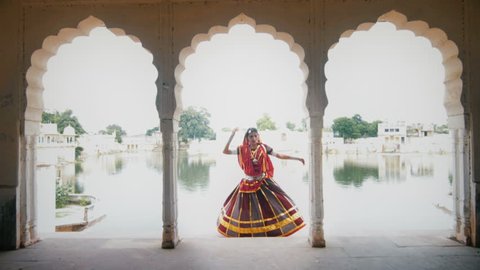 A wide shot of a graceful woman in traditional colorful cloths or attire performing in beautiful palace against the lake. An Indian elegant classical or folk dancer dancing in an ancient mansion