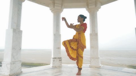 A wide shot of a confident Indian classical dancer dancing elegantly in white marble temple located on a hilltop. A moving shot of an attractive woman in traditional yellow sari performing folk dance