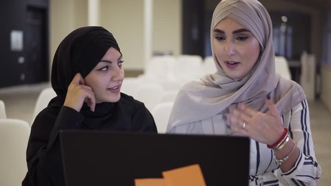 Pretty muslim businesswomen in hijab at office workplace or conference hall. Two smiling arabic woman working on laptop on startup project together, discussing the cases, typing on the laptop