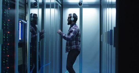 View of adult bearded man in headphones dancing in corridor of server room excited with finished workday
