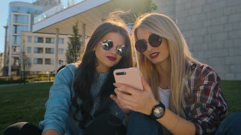 Portrait of two happy hipster girls in casual clothing relax and chat over. Woman using smart phone typing text message outside. They are happy and smiling, sitting on the grass