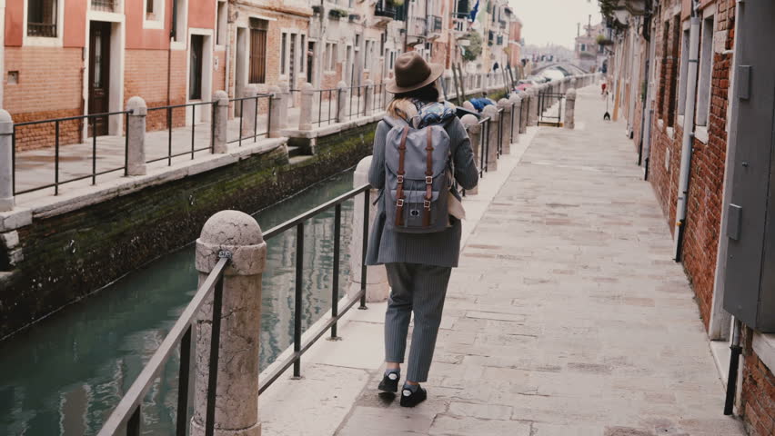 Happy young stylish European woman tourist with camera enjoying walking along famous old canal street in Venice Italy.