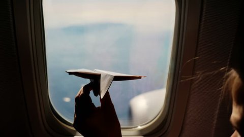 close-up. Silhouette of a child's hand with small paper plane against the background of airplane window. Child sitting by aircraft window and playing with little paper plane. during flight on airplane
