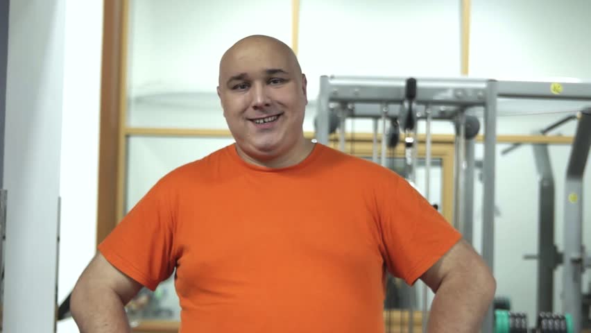 Fat handsome man in a gym in orange t-shirt standing and looking happily to the camera, smiling, healthcare motivation 