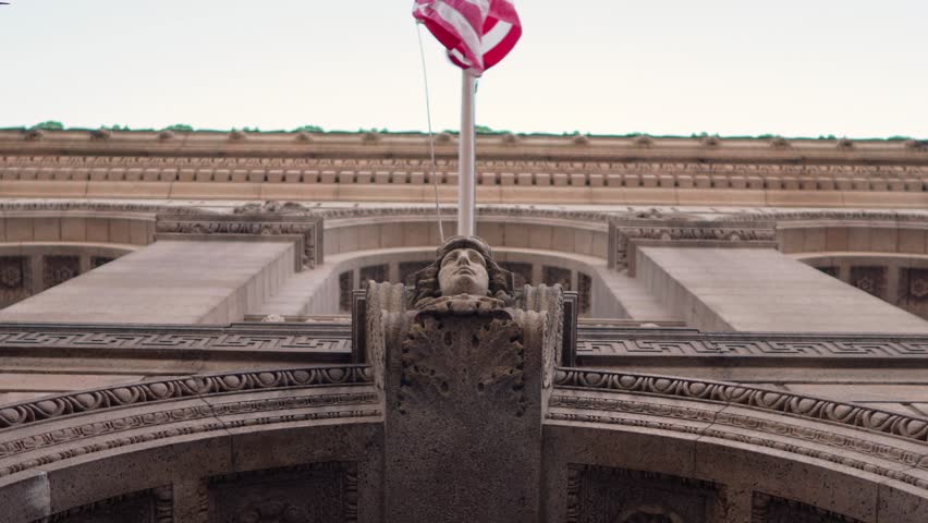 A bronze head looks toward Copley Square in front of the Boston Public Library in Boston, MA USA while an American flag flaps slowly with a lens flare.