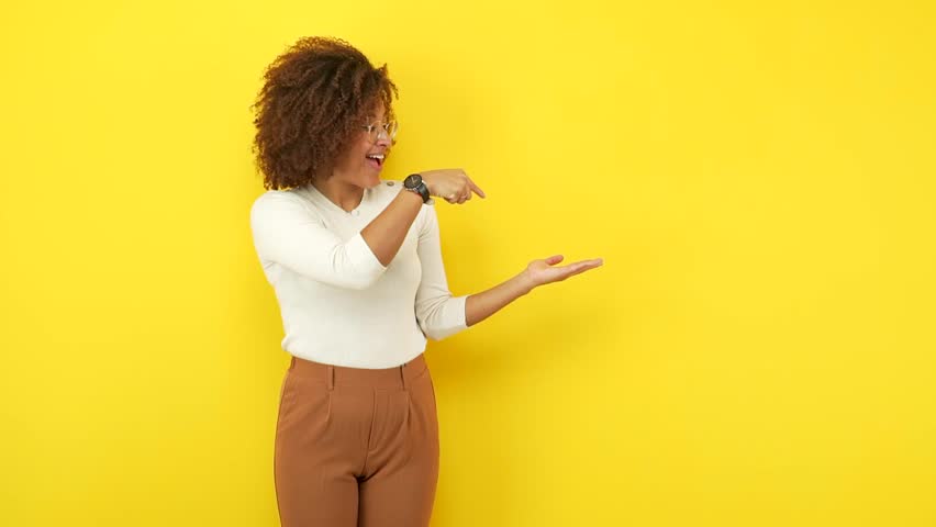 Young african american woman against a yellow wall holding something with hand, showing a product, smiling and cheerful, offering an imaginary object