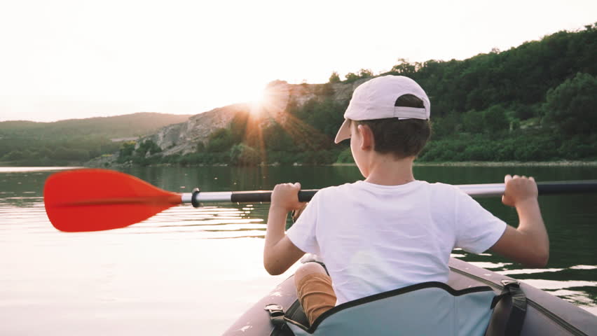 Happy boy with her dog Jack Russell breed paddling on an inflatable kayak on water of large mountain lake on background of beautiful orange sunset slow motion. Family sports holiday. Go Everywhere