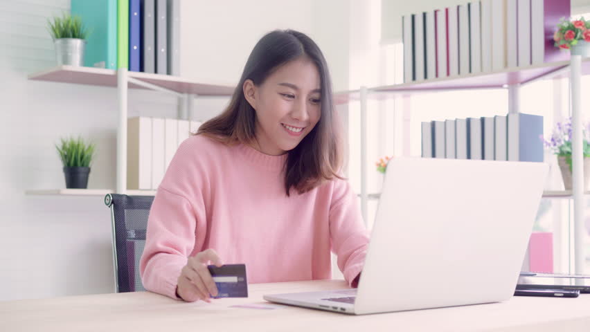 Beautiful Asian woman using computer or laptop buying online shopping by credit card while wear sweater sitting on desk in living room at home. Lifestyle woman at home concept. | Shutterstock HD Video #1018419298
