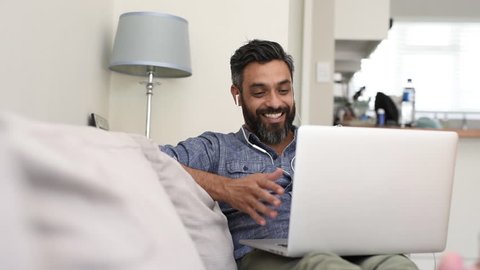Relaxed mature man using laptop with earphones for a video call. Cheerful smiling latin man sitting on couch having a friendly video call. Happy middle eastern man enjoying video calling on sofa.