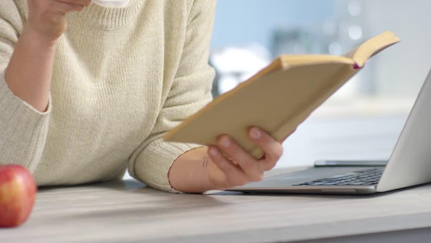Handheld tilt up of cheerful curly woman in round glasses smiling and drinking coffee while reading book in morning