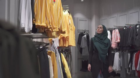 Young pretty muslim woman in a hijab and with a backpack chooses clothes in the store