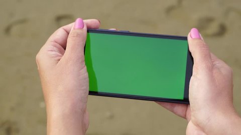 Closeup top view of modern cell phone in hands of european woman standing on sandy sea beach at hotel resort. Monitor of device with empty blank green screen. Real time 4k video footage.