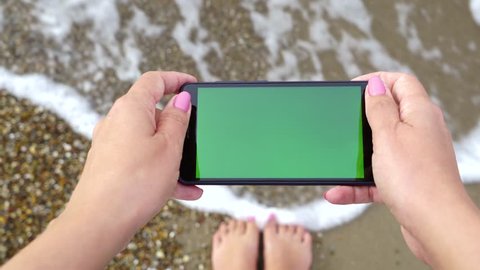 Closeup top view of modern cell phone in hands of happy woman standing on sandy sea beach at hotel resort. Monitor of device with empty blank green screen. Point of view real time 4k video footage.