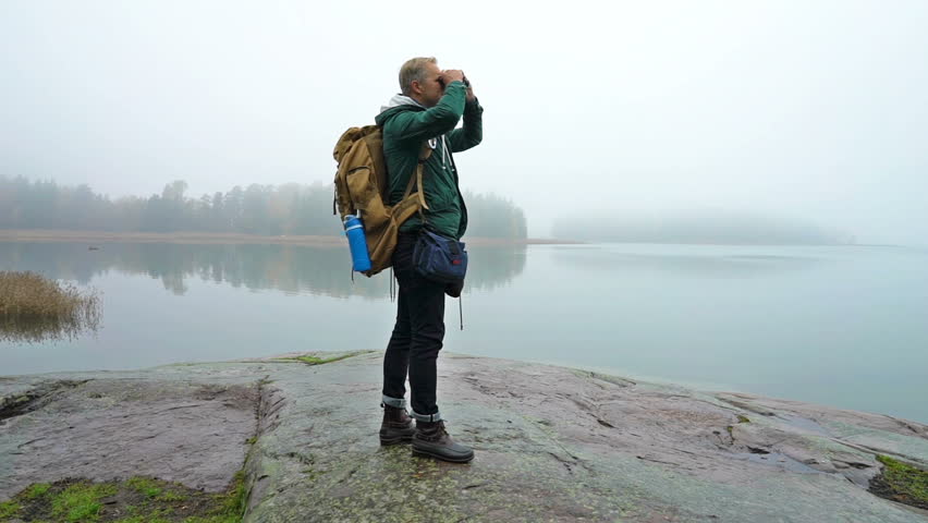 A middle-aged man walking on a rocky shore of the sea on a misty autumn morning and watching wildlife through binoculars. Slow motion
