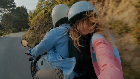 Young attractive couple of fashionable hipsters or millennials driving towards new exciting travel destination on motorbike on mountain forest road, wear white helmets, girl makes selfie.April 2018