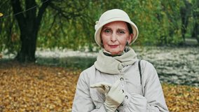Fashionable elderly lady in an elegant autumn outfit standing outdoors in a park looking thoughtfully at the camera as in pans around her